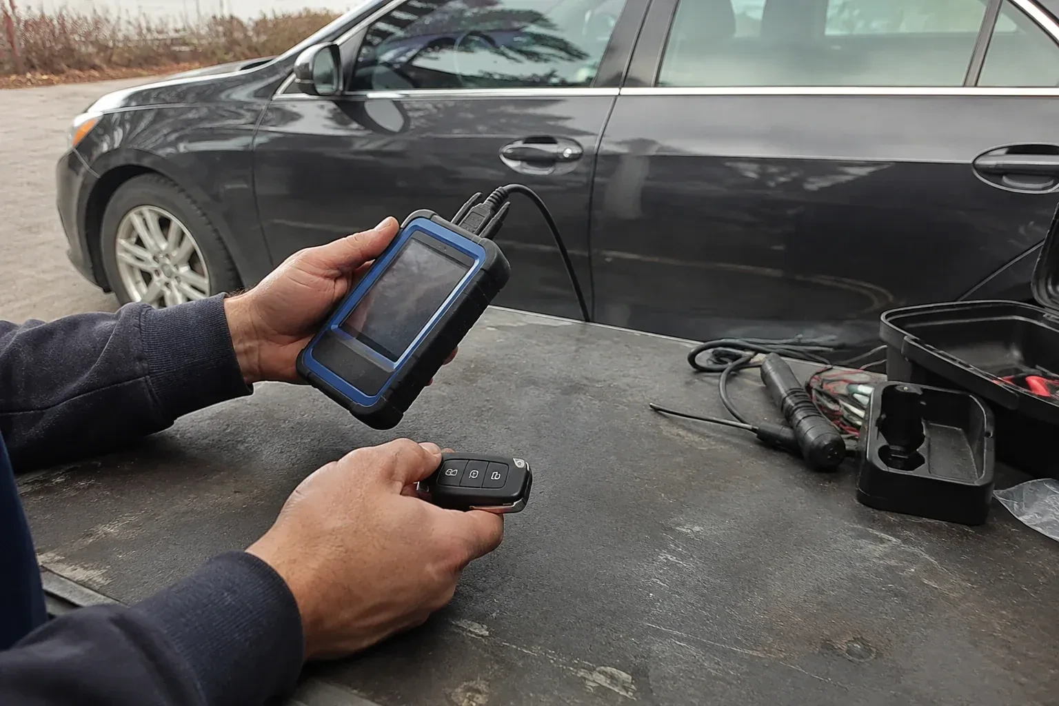 Hands holding a blue-edged electronic device and car-key fob at a worktable beside a parked car. — Transponder Key Programming, Caledon